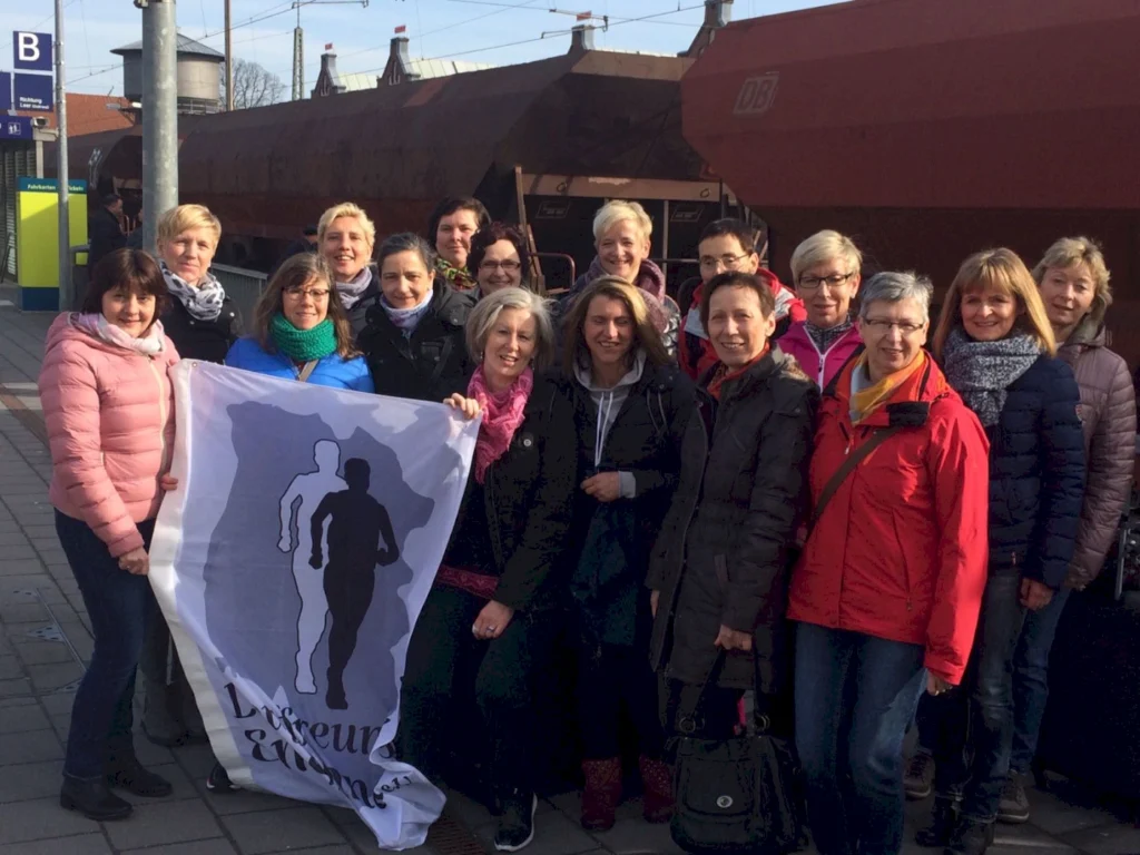 Gruppe von Frauen, die mit einer Flagge der Laufgemeinschaft posieren, vor einem Bahnhof in Deutschland, umgeben von einem Güterwagen im Hintergrund.