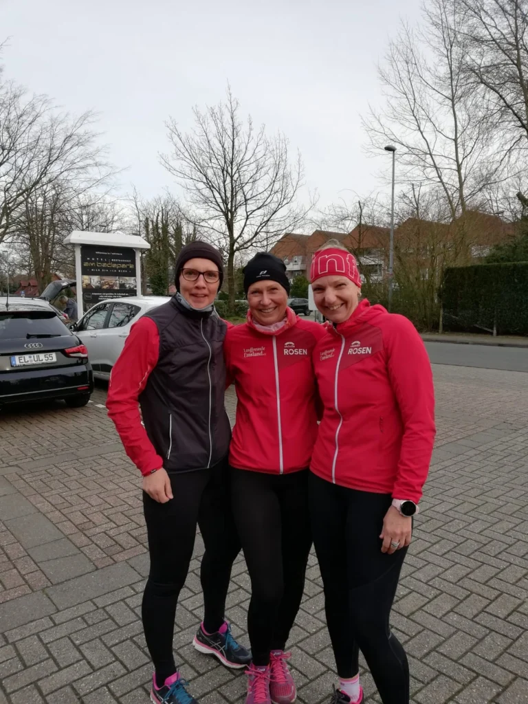 Drei sportlich gekleidete Frauen posieren lächelnd in Laufbekleidung in einer Stadtansicht, umgeben von Bäumen und parkenden Autos. Sie tragen rote Jacken mit dem Logo "Rosen" und sind bereit für einen Lauf oder ein Training.