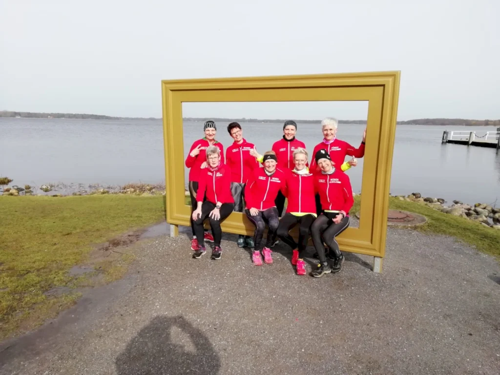 Gruppe von acht Frauen in roten Sportjacken posiert vor einem goldenen Bilderrahmen am Ufer eines Gewässers, umgeben von einer grünen Wiese und Steinchen.
