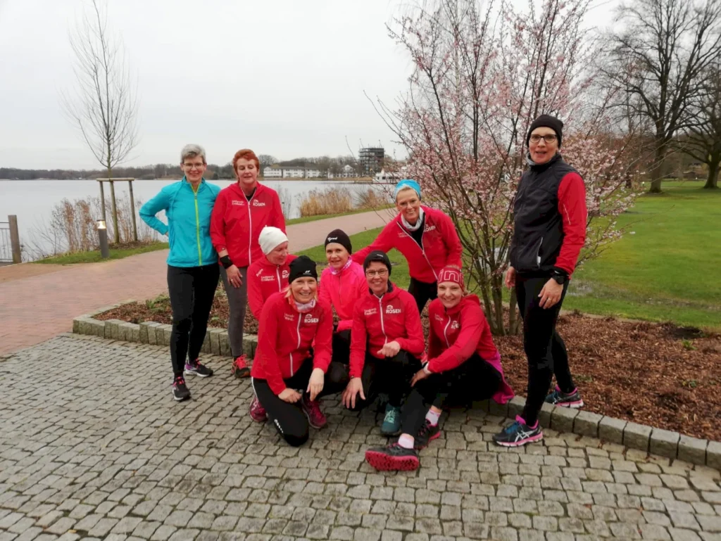 Gruppe von Läuferinnen in roten Jacken posiert am Ufer eines Sees, umgeben von blühenden Bäumen und einer gepflasterten Fläche. Ideal für einen Lauf- oder Sportevent.