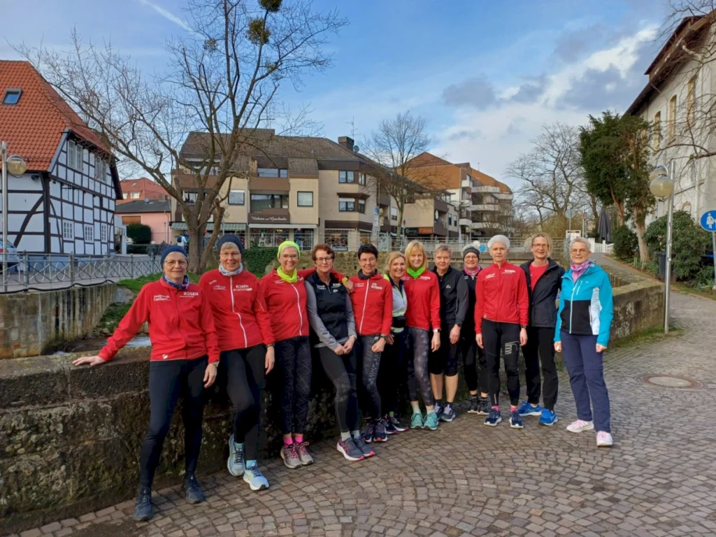 Gruppe von Läuferinnen in roten Trainingsjacken, die an einer Steinmauer in einer Stadt stehen. Im Hintergrund sind traditionelle Fachwerkhäuser und moderne Gebäude zu sehen. Der Himmel ist blau mit einigen Wolken.