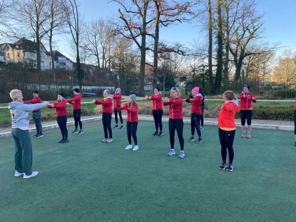 Gruppe von Sportlern in roten Jacken beim Outdoor-Training in einem Park, die verschiedene Übungen durchführen und von einer Trainerin angeleitet werden.