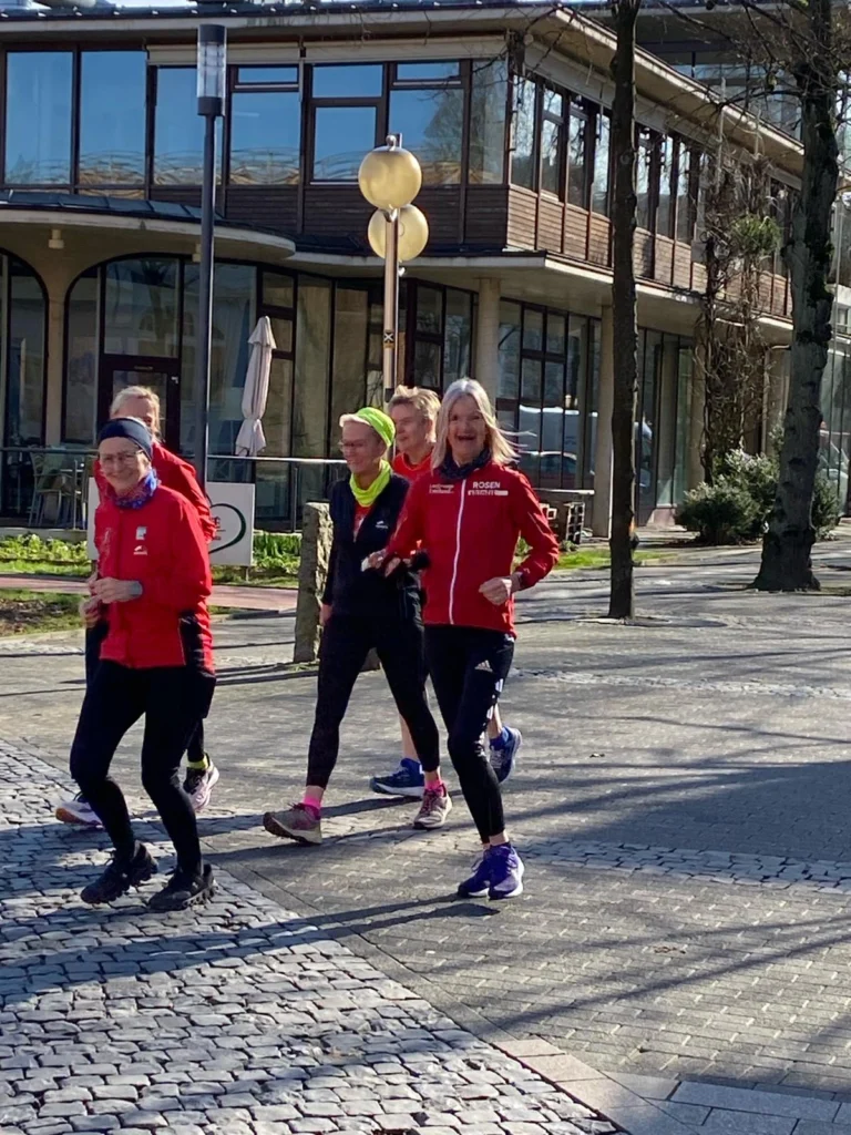 Gruppe von Läuferinnen in roten Jacken, die auf einem gepflasterten Weg joggen, umgeben von modernen Gebäuden und Bäumen, bei sonnigem Wetter.