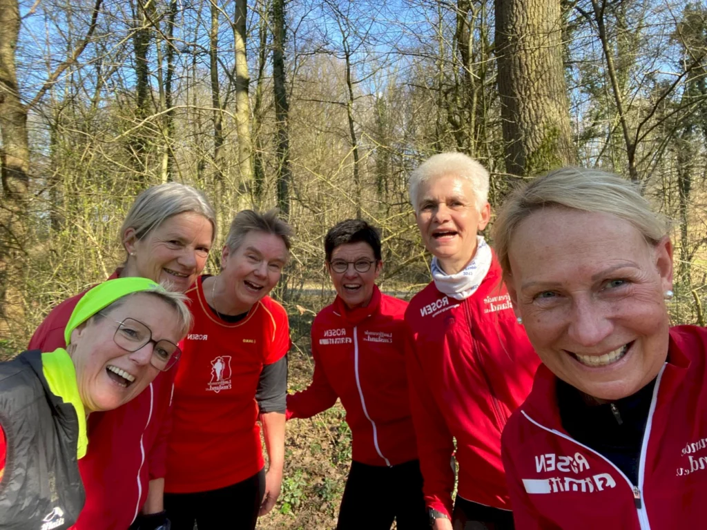 Gruppe von sechs Frauen in roten Laufjacken, die in einem Wald lächeln und Spaß haben, während sie eine gemeinsame Aktivität im Freien genießen.