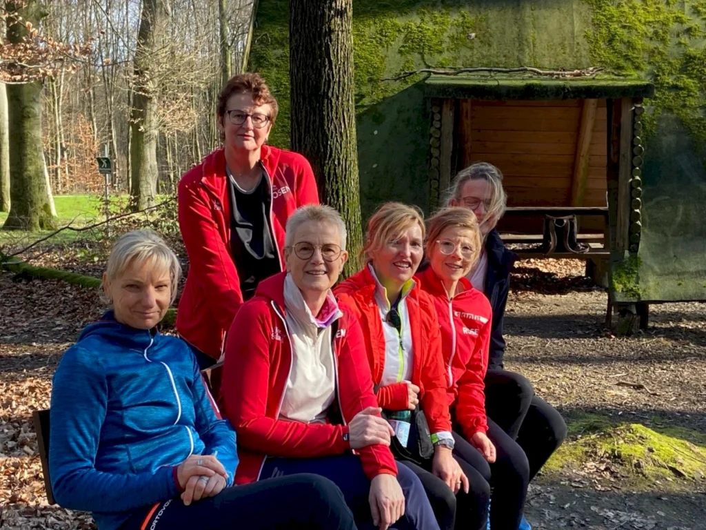 Sechs Frauen in sportlicher Kleidung sitzen in einem Waldgebiet und genießen die Natur. Sie lächeln in die Kamera, während im Hintergrund eine rustikale Holzhütte und Bäume zu sehen sind. Ideal für Themen wie Outdoor-Aktivitäten, Fitness und Gemeinschaft.