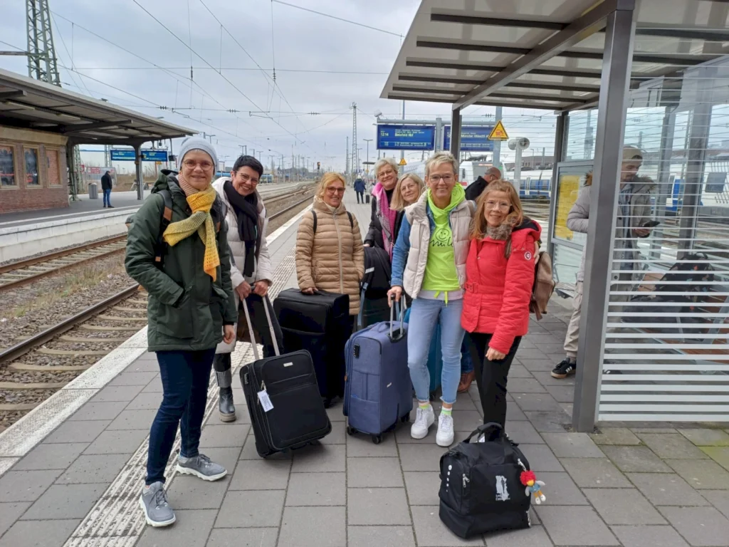 Gruppe von Reisenden am Bahnhof mit Gepäck, bereit für die Abreise. Sie stehen auf dem Bahnsteig, umgeben von Zügen und Bahnschildern, während sie auf ihren Zug warten. Ideal für Reise- und Transportthemen.