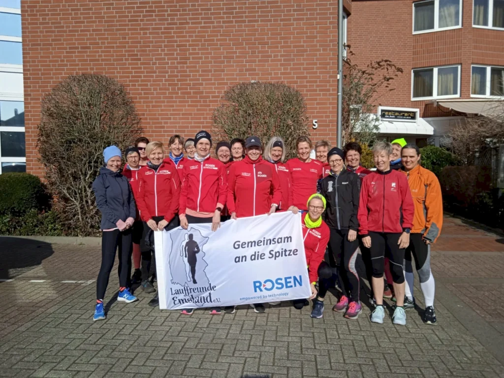 Gruppe von Laufbegeisterten in roten Jacken, die vor einem Gebäude stehen und ein Banner mit der Aufschrift „Gemeinsam an die Spitze“ halten. Die Teilnehmerinnen zeigen Freude und Begeisterung für den Laufsport, während sie sich für ein gemeinsames Training versammeln.