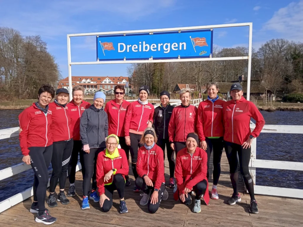 Gruppe von Frauen in roten Laufjacken auf einem Steg am Wasser in Dreibergen, umgeben von Bäumen und einem blauen Himmel, während sie für ein Gruppenfoto posieren.