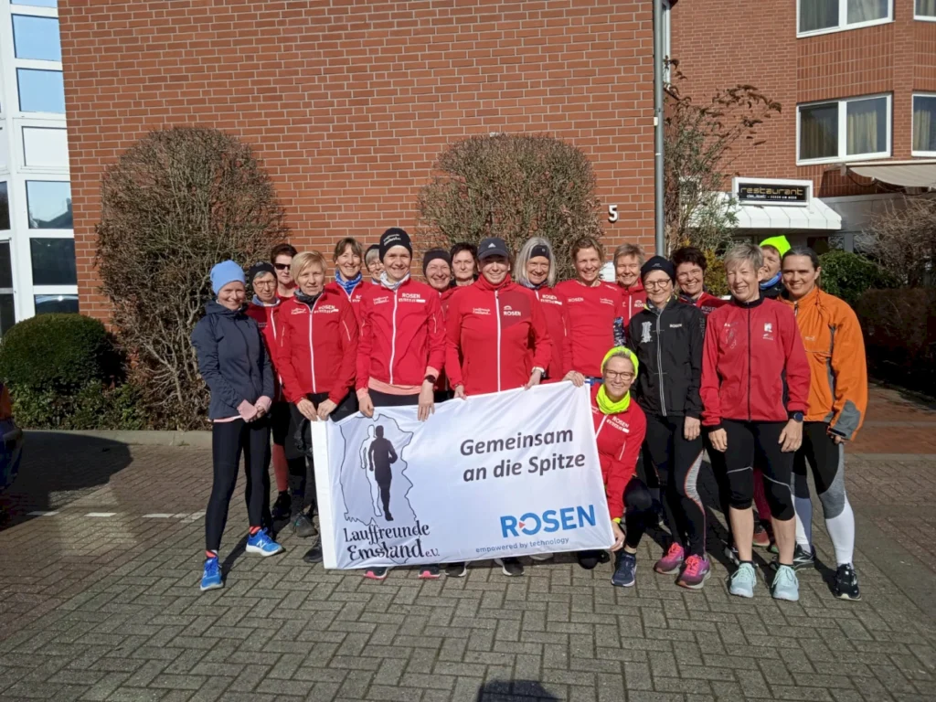 Gruppenfoto einer Laufgemeinschaft in roten Jacken vor einem Gebäude, mit einem Banner, das die Botschaft "Gemeinsam an die Spitze" zeigt, symbolisiert Teamgeist und sportliche Aktivität.