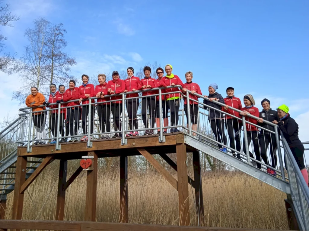 Gruppe von 16 Frauen in roten Trainingsjacken auf einer Aussichtsplattform, umgeben von Natur und blauem Himmel, während sie lächeln und posieren. Ideal für Themen wie Sport, Fitness und Gemeinschaft.