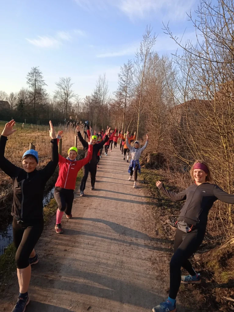 Gruppe von Läufern in Sportkleidung, die auf einem Weg im Freien stehen und mit erhobenen Händen Freude zeigen. Umgeben von Bäumen und einem klaren Himmel, vermittelt das Bild eine positive und motivierende Atmosphäre für das Laufen im Freien.