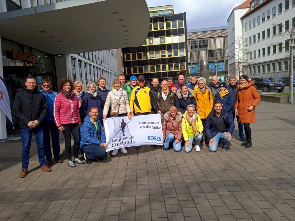 Gruppenfoto von Mitgliedern der Laufgruppe Emstland e.V. vor dem InterCity Hotel, die gemeinsam für den Sport an die Spitze stehen. Die Teilnehmer tragen bunte Sportbekleidung und halten ein Banner mit der Aufschrift "Gemeinsam an die Spitze".