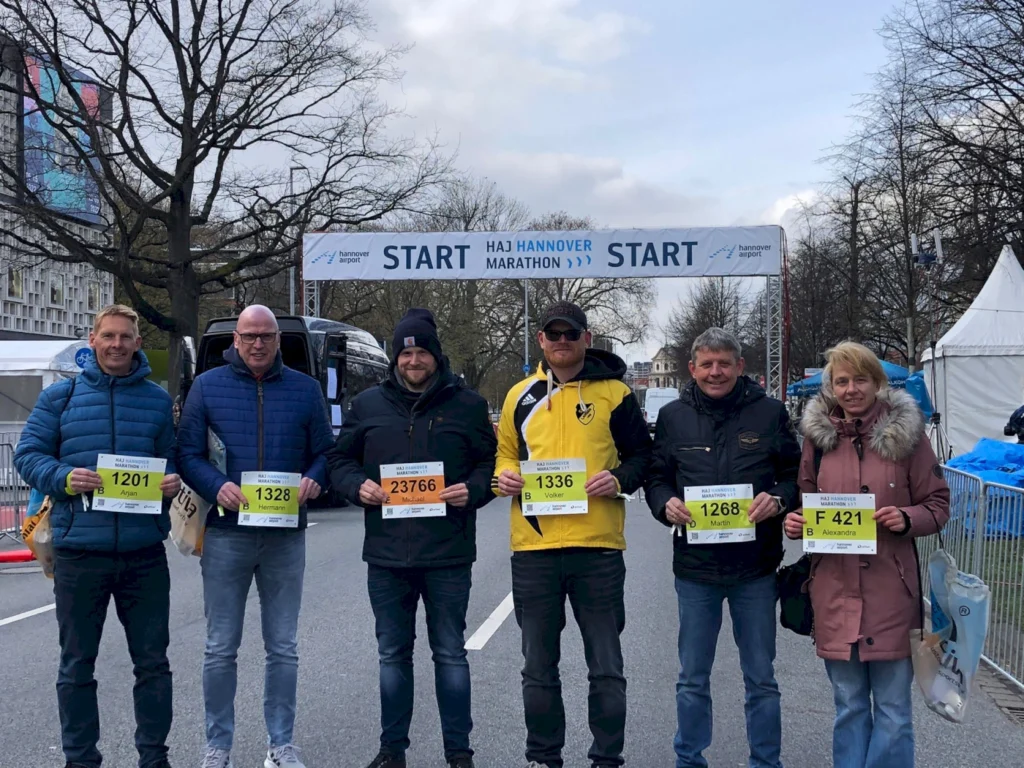 Sechs Teilnehmer stehen am Start des HAJ Hannover Marathons, jeder mit einer Startnummer in der Hand. Die Szene zeigt eine frische, bewölkte Atmosphäre, während die Teilnehmer in Sportkleidung posieren. Im Hintergrund ist das Startbanner deutlich sichtbar, das den Beginn des Marathons ankündigt.