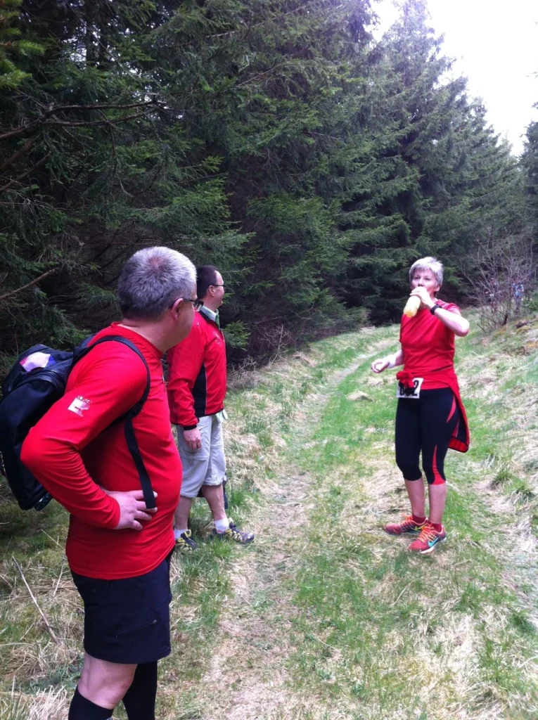 Drei Personen in roten T-Shirts stehen auf einem Waldweg, umgeben von hohen Bäumen. Eine Person trinkt aus einer Flasche, während die anderen in die Ferne schauen. Die Szene vermittelt eine aktive Outdoor-Atmosphäre und zeigt eine Gruppe von Wanderern oder Läufern in der Natur.