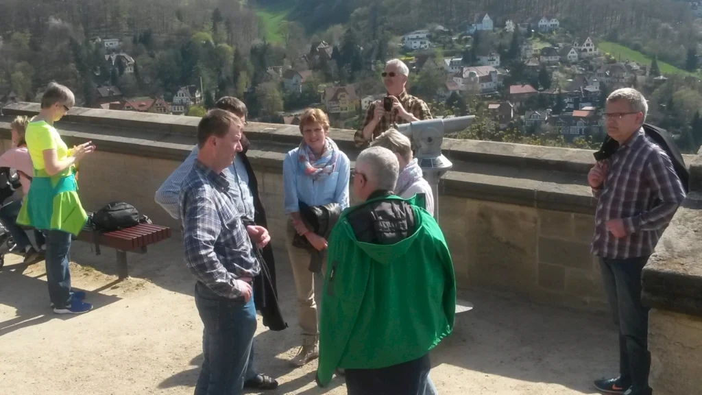 Gruppe von Touristen, die an einem Aussichtspunkt stehen und die Landschaft betrachten, mit Blick auf eine malerische Stadt im Hintergrund. Ein Mann in einem grünen Jacket spricht mit anderen, während eine Frau in einem leuchtend gelben T-Shirt ihr Handy benutzt.