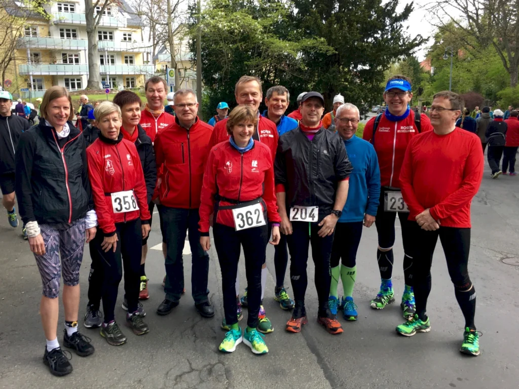 Gruppe von Läufern in roten Trikots, die an einem Wettlauf teilnehmen, stehen zusammen auf der Straße. Einige tragen Startnummern, während im Hintergrund andere Teilnehmer vorbeilaufen. Die Szene zeigt eine aktive Laufveranstaltung in einer städtischen Umgebung.