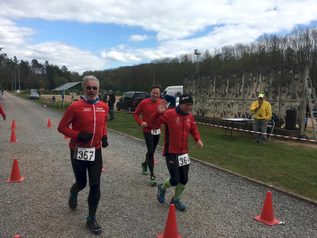 Drei Läufer in roten Trikots nehmen an einem Wettlauf teil, während sie auf einer Schotterstrecke joggen. Im Hintergrund sind Zuschauer und eine Sportanlage zu sehen.