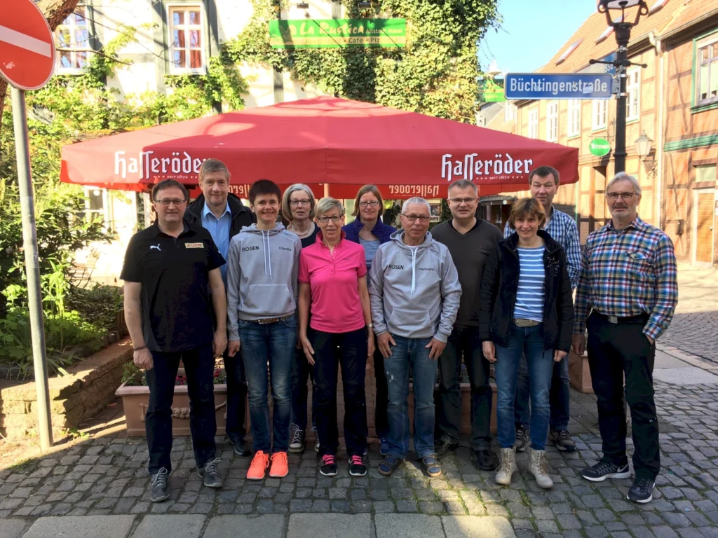 Gruppe von Menschen vor einem Restaurant mit rotem Sonnenschirm in einer malerischen Straße, umgeben von historischen Gebäuden und Straßenschildern.