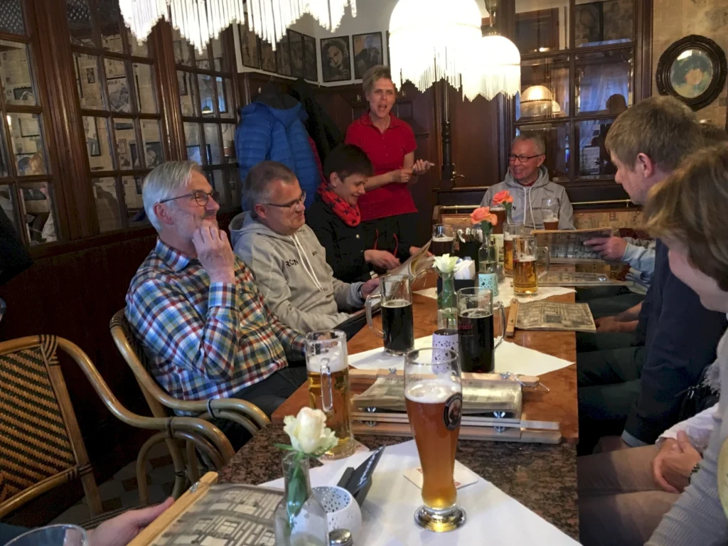 Gesellige Runde in einem traditionellen Restaurant mit mehreren Personen, die an einem Tisch sitzen, Essen und Getränke genießen. Im Hintergrund ist eine Kellnerin zu sehen, während die Gäste sich unterhalten und die Speisekarten studieren. Die Atmosphäre ist freundlich und einladend, mit dekorativen Elementen und Blumen auf dem Tisch.