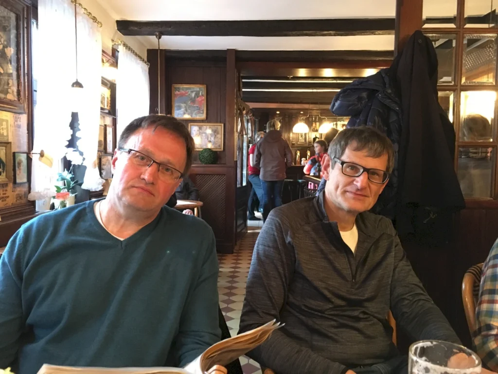 Zwei Männer sitzen an einem Tisch in einem gemütlichen Restaurant mit rustikaler Einrichtung. Der Mann links trägt einen blauen Pullover und sieht nachdenklich aus, während der Mann rechts in einem grauen Sweatshirt lächelt. Im Hintergrund sind weitere Gäste und eine einladende Atmosphäre zu sehen.