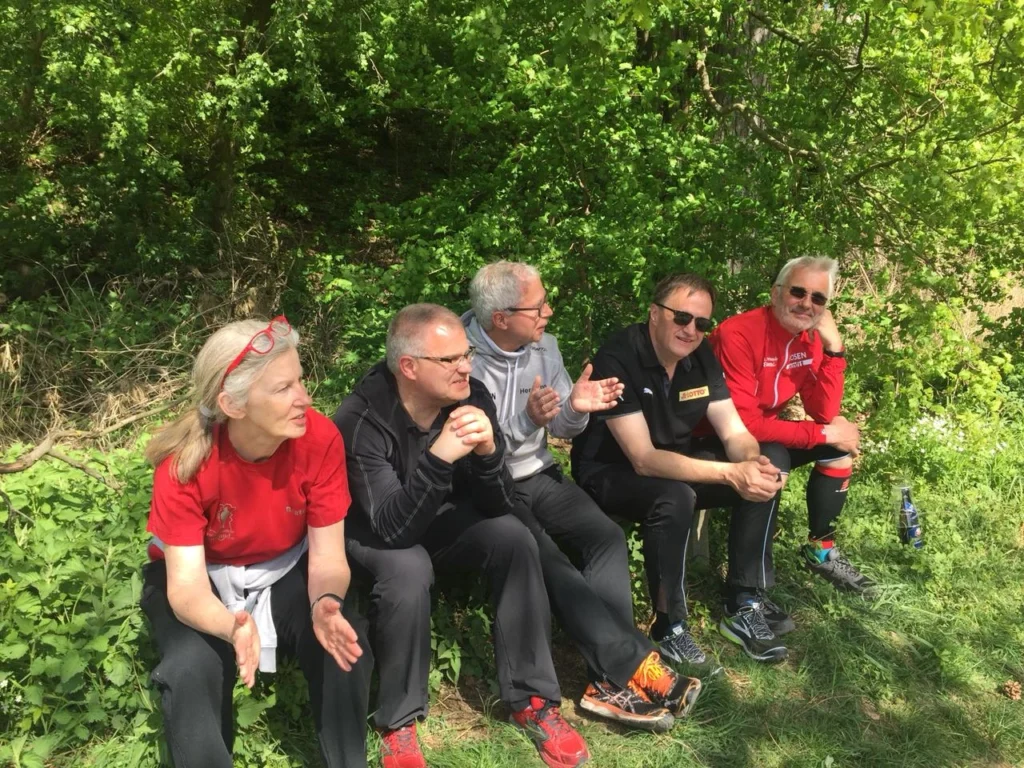 Gruppe von fünf Personen, die im Freien auf einer Wiese sitzen und sich unterhalten, umgeben von üppigem Grün und Bäumen. Ideal für Themen wie Teamarbeit, Outdoor-Aktivitäten und soziale Interaktionen.