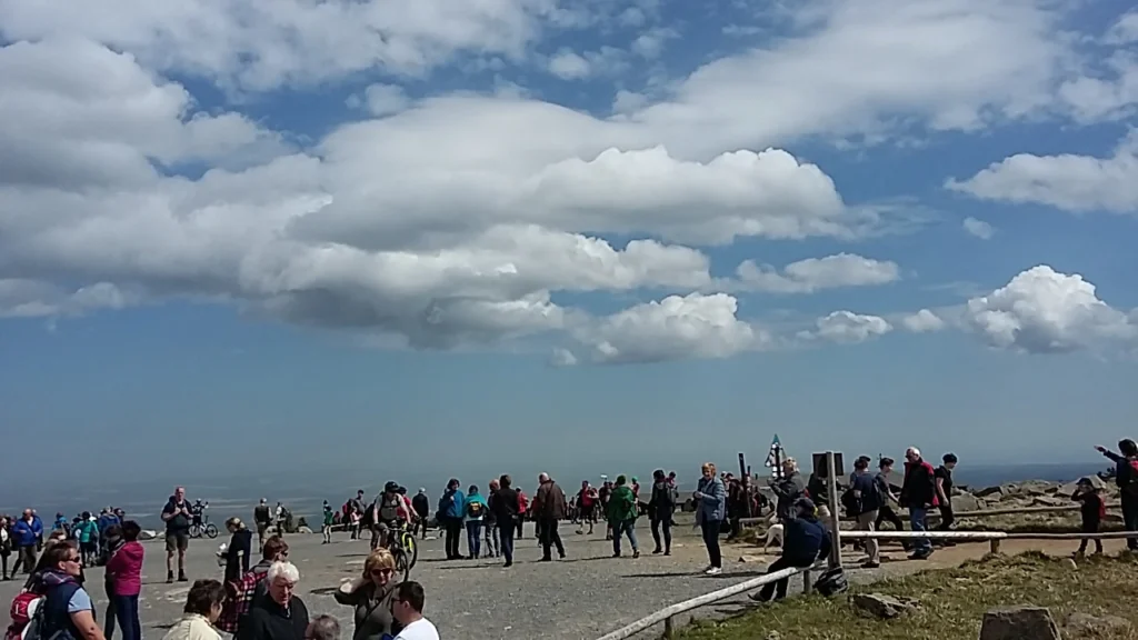 Menschenmenge auf dem Gipfel eines Berges, bei schönem Wetter mit blauen Himmel und Wolken, ideal für Wanderer und Touristen.