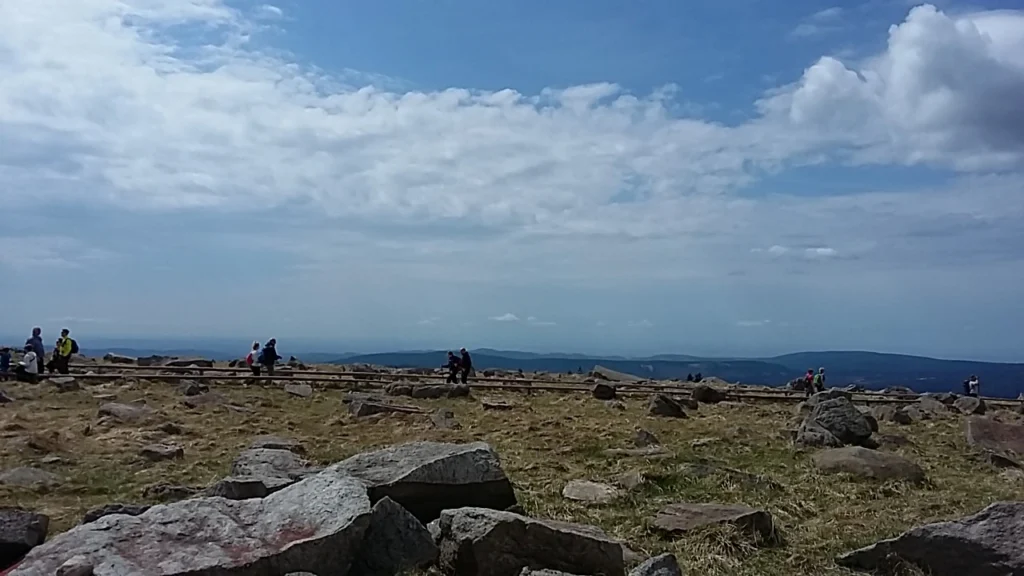 Panoramablick auf eine felsige Landschaft mit Wanderern auf einem Berggipfel, umgeben von Wolken und einer weiten Aussicht auf das Tal und die umliegenden Berge.