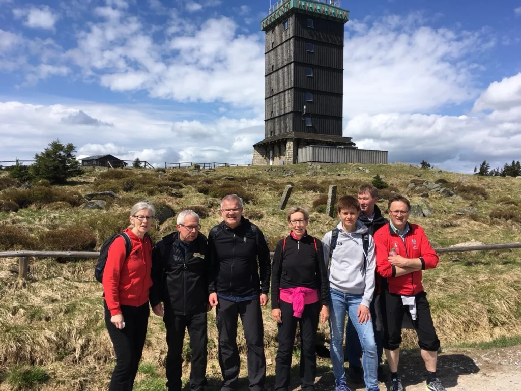 Gruppe von Wanderern vor einem Aussichtsturm in der Natur, umgeben von hügeliger Landschaft und blauem Himmel mit Wolken. Ideal für Outdoor-Aktivitäten und Erkundungen.