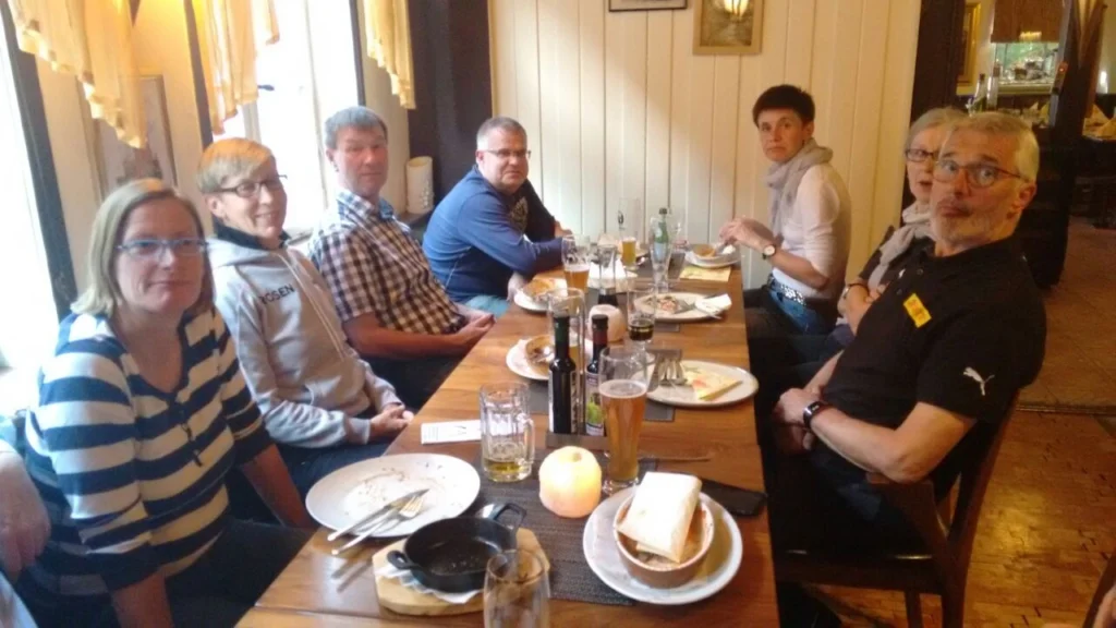 Gruppenszene in einem Restaurant mit sechs Personen, die an einem Tisch sitzen. Es gibt leere Teller, Gläser und Getränke auf dem Tisch. Die Atmosphäre wirkt gesellig und entspannt.