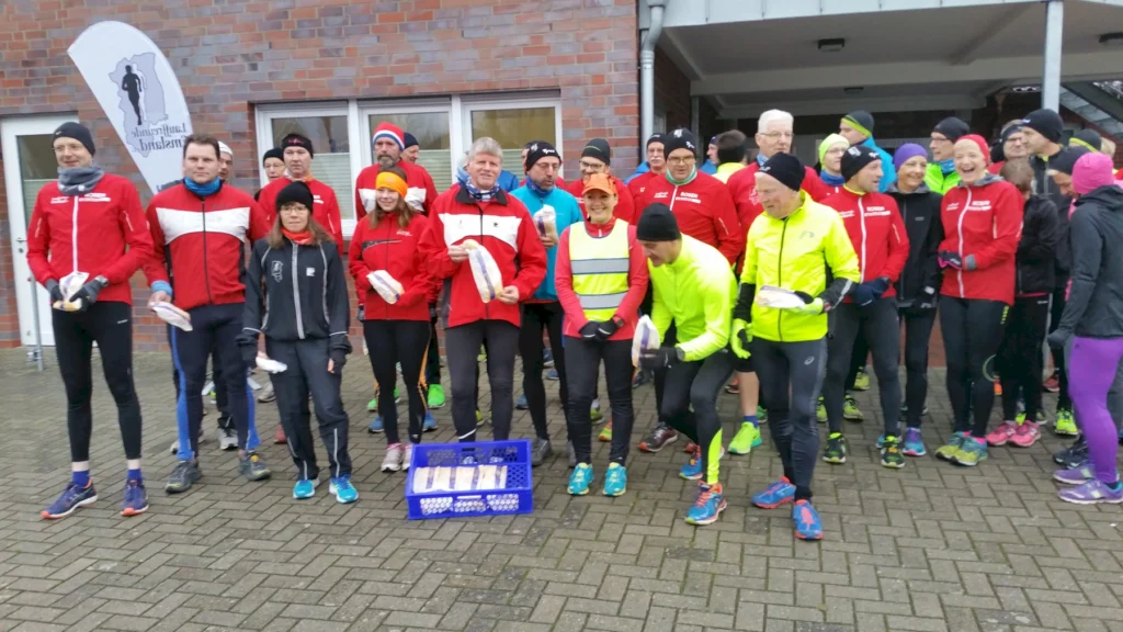 Läufergruppe in bunter Sportkleidung mit Brotpaketen beim Lauf-Event vor Backstein-Gebäude versammelt.