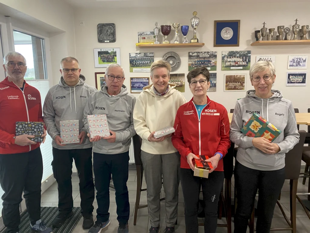 Gruppe von sechs Personen mit Geschenken in Vereinskleidung posiert in einem Raum mit Pokalen und Mannschaftsfotos an der Wand.