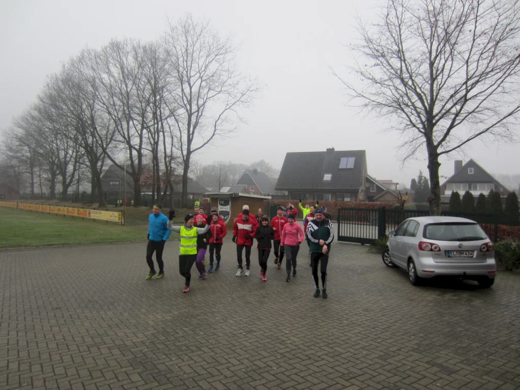 Gruppe von Menschen joggt im Winter durch ein Wohngebiet, Bäume ohne Laub und parkendes Auto sichtbar.
