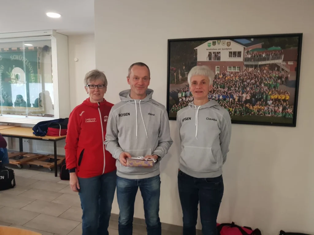 Drei Personen stehen vor einem Vereinsfoto in einem Raum und lächeln, eine hält eine Trophäe in der Hand.