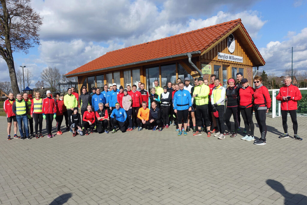 Gruppe von Läufern in bunter Sportkleidung steht vor einem Vereinsgebäude mit roten Dachziegeln und Willkommen-Schild.