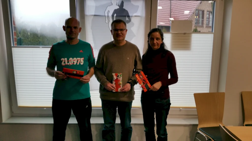 Drei Personen mit Geschenken vor einem Fenster, im Hintergrund eine Silhouette von Läufern.