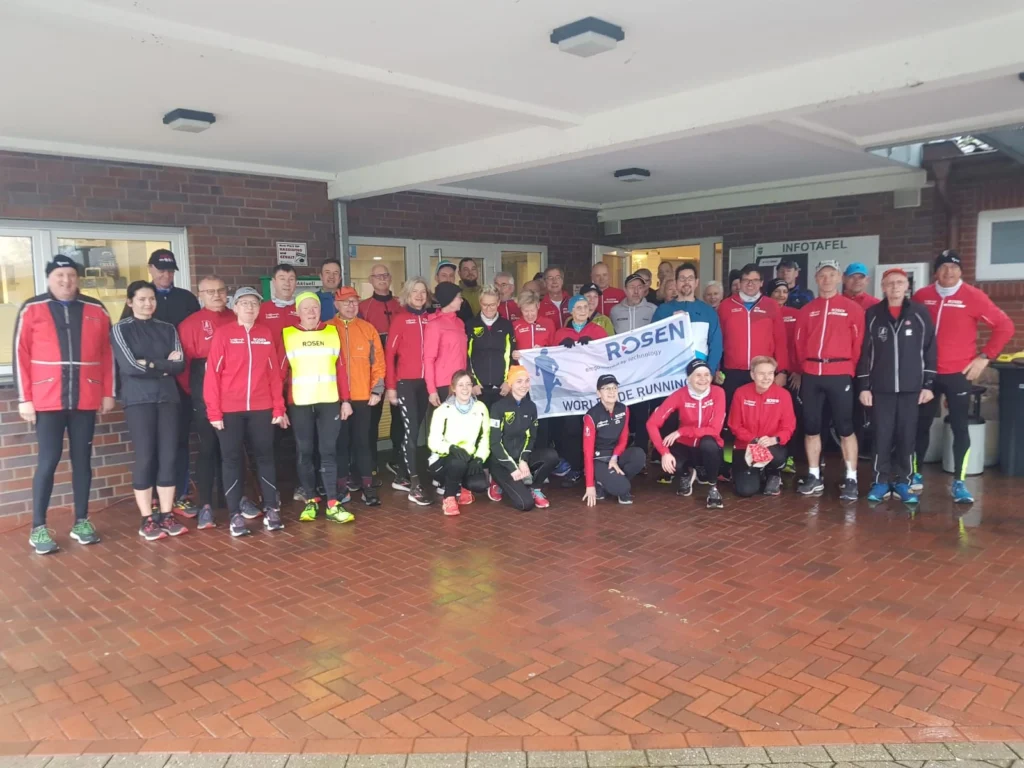 Gruppe von Läufern in Sportkleidung versammelt für ein Event, einige halten ein Banner mit der Aufschrift “Rosen”.