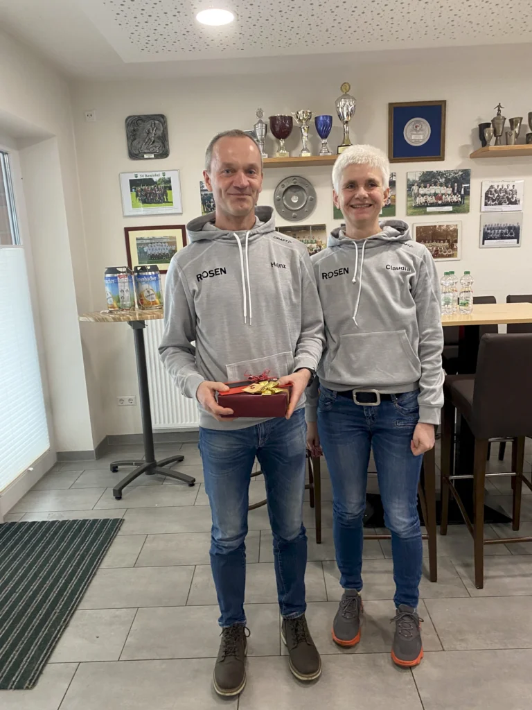 Zwei Personen in grauen Pullovern stehen lächelnd in einem Raum mit Pokalen und Erinnerungsfotos.