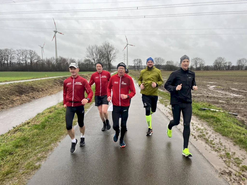 Gruppe von Joggern läuft bei bewölktem Wetter auf Landstraße, Windräder im Hintergrund, winterlich gekleidet.