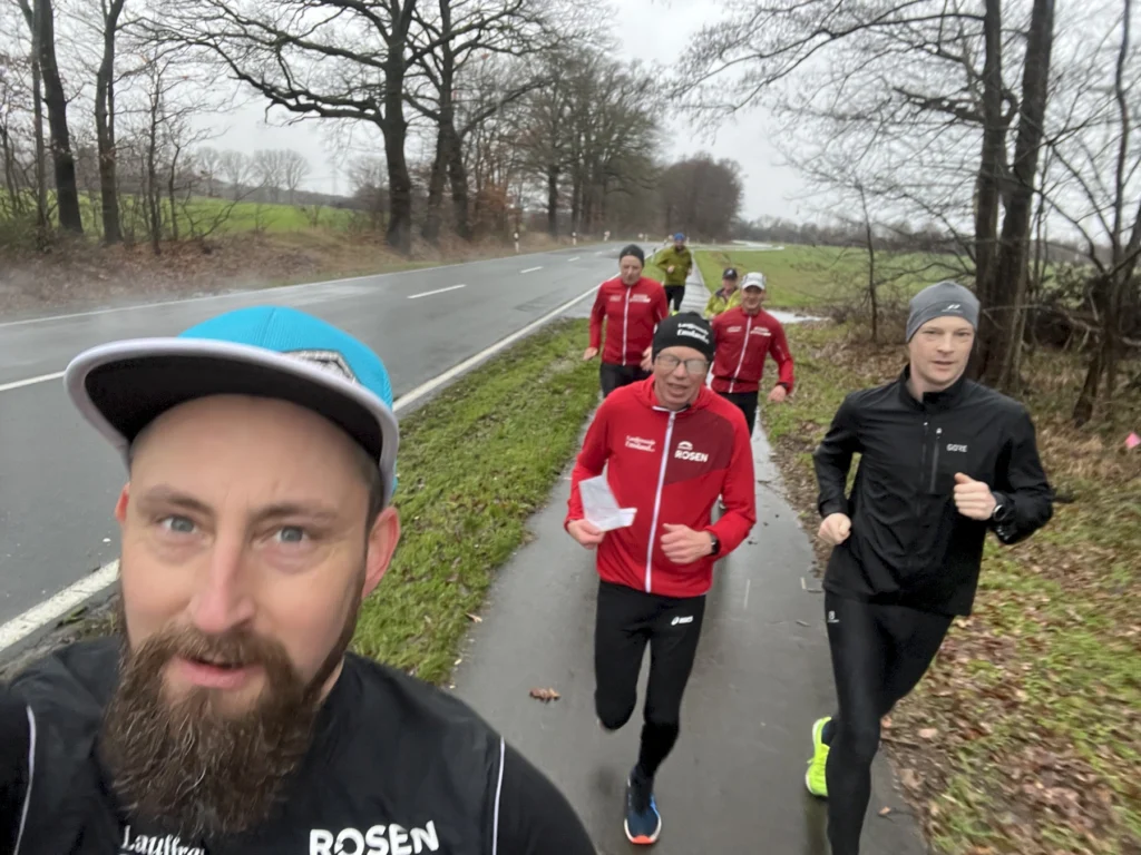 Läufergruppe joggt bei grauem Wetter auf einem ländlichen Weg entlang einer Straße, mit Bäumen im Hintergrund.