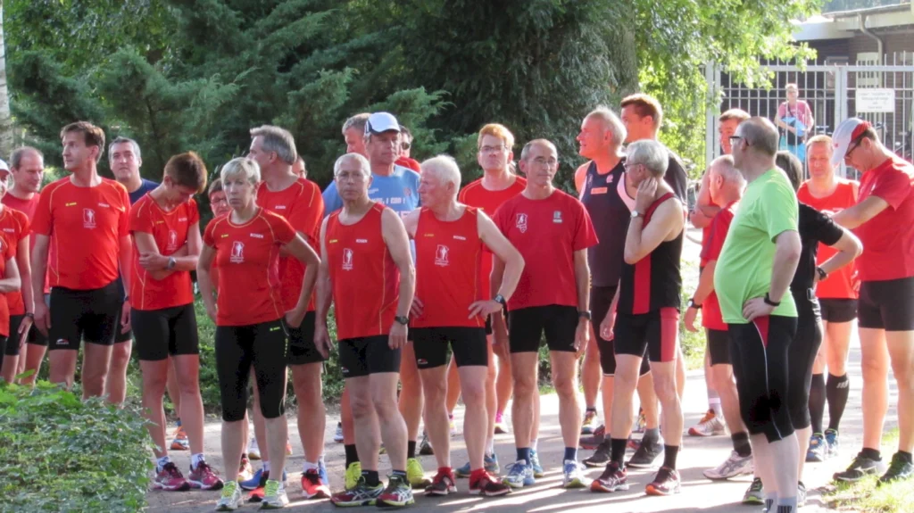 Gruppierung von Läufern in roten Trikots, die sich vor einem Wettkampf versammeln, umgeben von Bäumen und einer Sporteinrichtung.