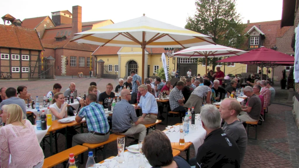 Menschen sitzen an langen Tischen unter Sonnenschirmen in einer geselligen Atmosphäre, während sie Speisen und Getränke genießen. Im Hintergrund sind historische Gebäude und eine gepflasterte Fläche zu sehen, die eine einladende Umgebung für ein Fest oder eine Veranstaltung schaffen.