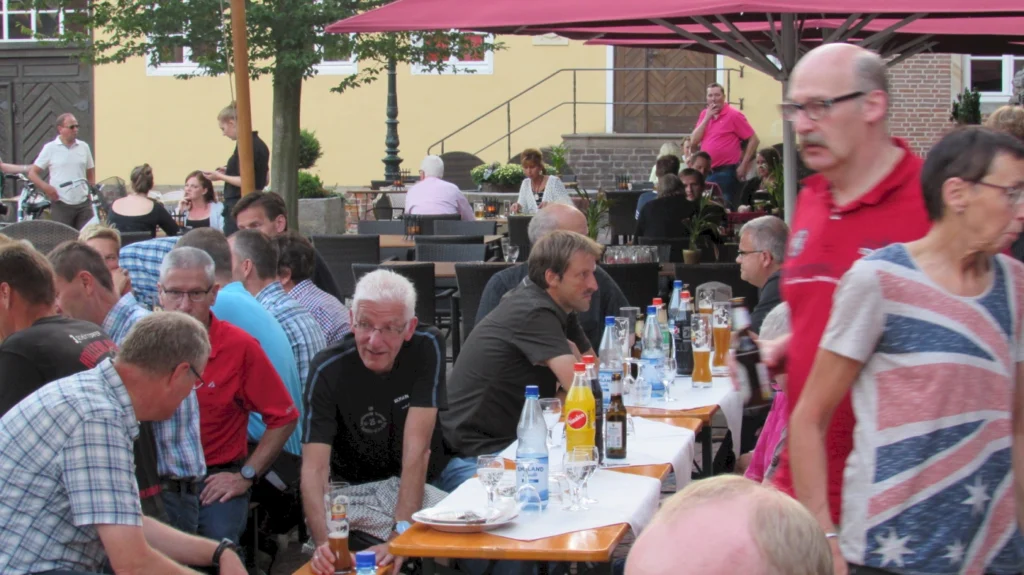 Belebte Außenterrasse eines Restaurants mit Gästen, die an Tischen sitzen, Getränke und Essen genießen. Im Hintergrund sind weitere Gäste und ein Kellner zu sehen. Die Atmosphäre ist gesellig und entspannt.