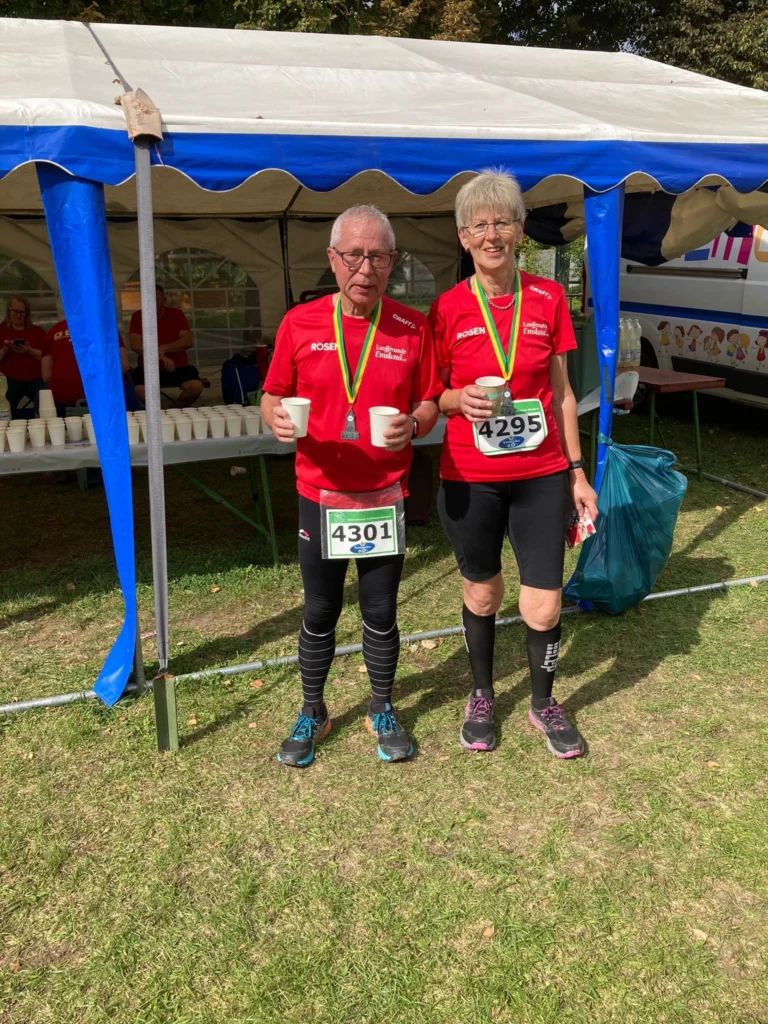 Zwei Seniorläufer mit Medaillen und Startnummern nach einem Rennen vor einem Zelt, halten Becher in der Hand.