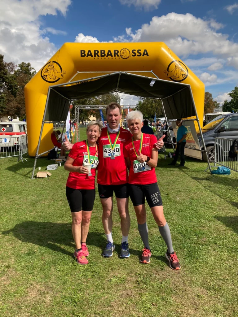 Drei Läufer in roten Trikots posieren lächelnd nach einem Wettkampf unter einem gelben Torbogen mit „Barbarossa“-Logo.