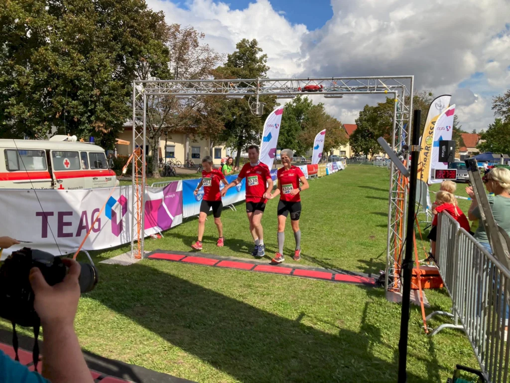 Mehrere Läufer überqueren gemeinsam die Ziellinie eines Rennens im Freien an einem sonnigen Tag.