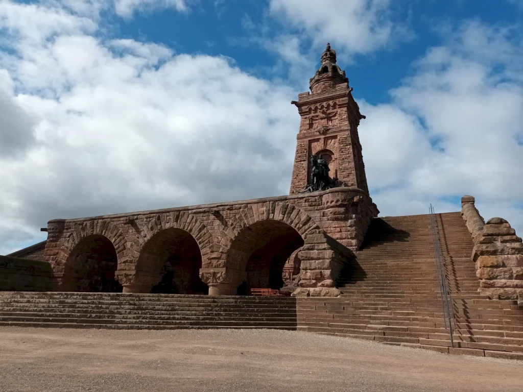 Monumentales Torbogen-Bauwerk aus Stein mit Treppe, blauem Himmel und Wolken im Hintergrund.