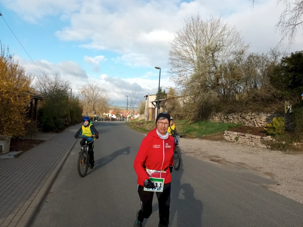 Jogger und zwei Radfahrer auf einer Dorfstraße an einem sonnigen Tag. Bäume ohne Blätter im Hintergrund.