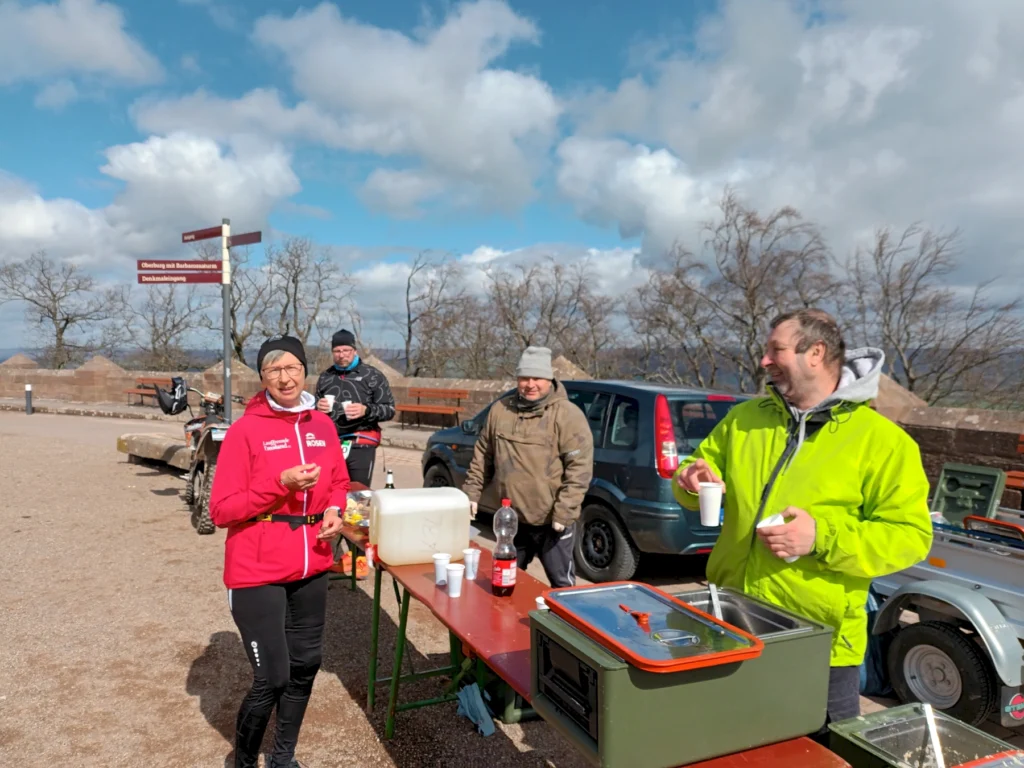 Gruppe von Menschen genießt eine Pause an einem Rastplatz, mit Getränken und Snacktisch im Freien bei sonnigem Wetter.