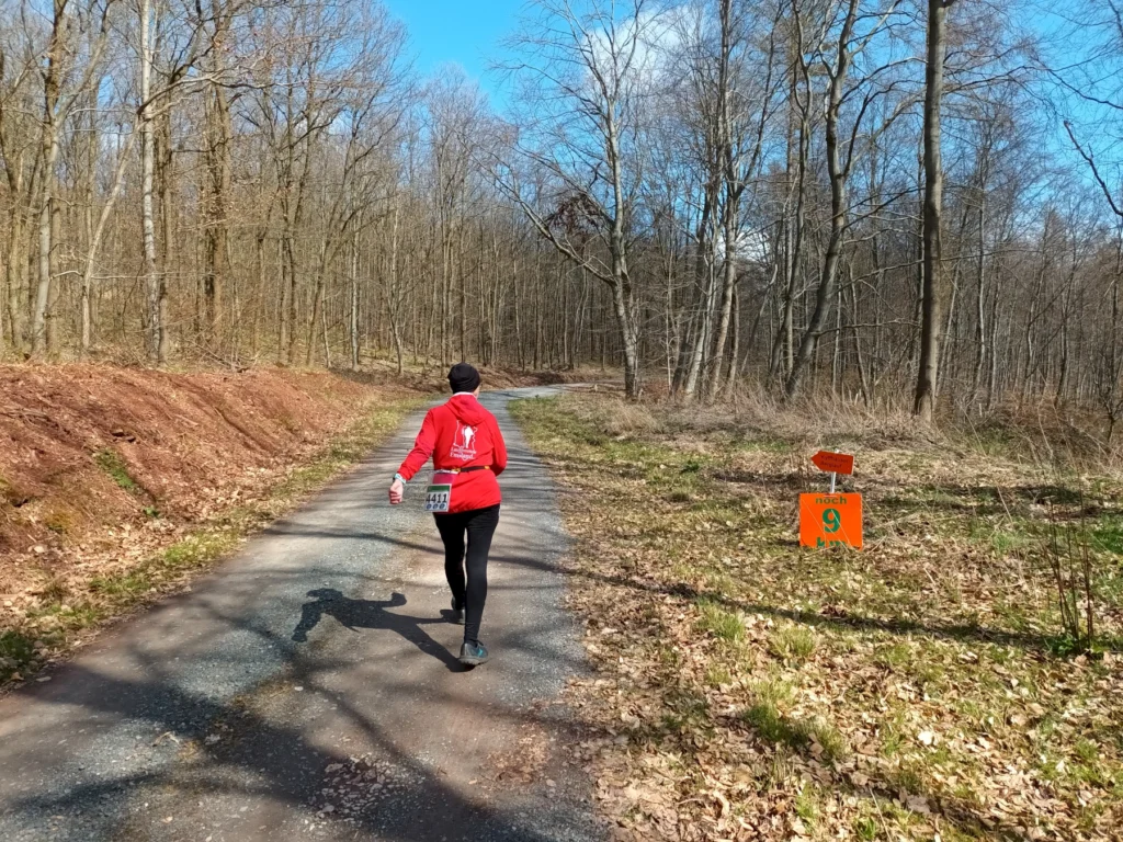 Läuferin im roten Shirt auf Waldweg, noch 9 km Schild. Klarer Himmel, Frühling. Wettkampf- oder Trainingslauf.