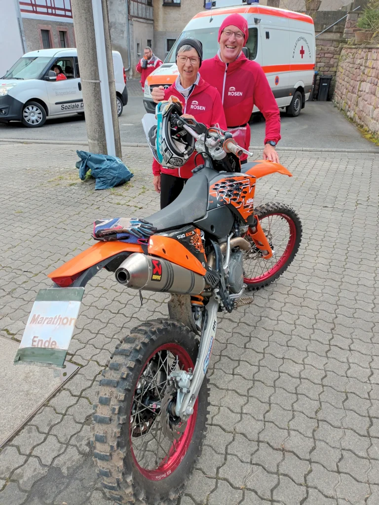 Zwei Personen in roten Jacken stehen neben einem orangefarbenen Motorrad, im Hintergrund ein Rettungsfahrzeug.